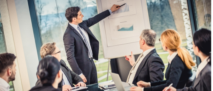 A businessman stands at a whiteboard with a graph addressing seated colleagues