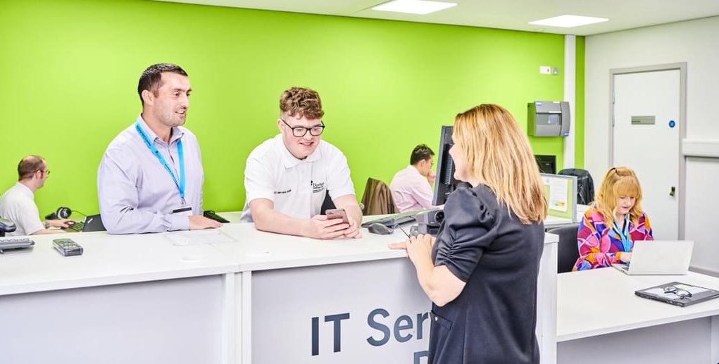 Long shot of an interior - it's a modern space - mainly pale grey with one fluorescent green wall. A young man in a white polo shirt sits behind a counter. Behind him are people working at monitors. An older man in a smart shirt and a blue lanyard stands nearby. A woman in a black dress stands on the other side of the counter. She has her back to camera. They are in the CIS help-desk section based