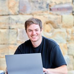 Student in castle courtyard picnic benches smiling at camera. He is wearing a black t-shirt and has brown hair. He is working on a laption