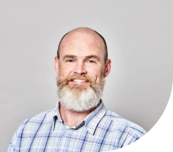 A photo of a man with a full beard and a checked shirt smiling at the camera