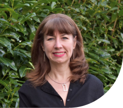 A photo of a woman with shoulder length brown hair and a fringe wearing a dark shirt, standing in front of a hedge