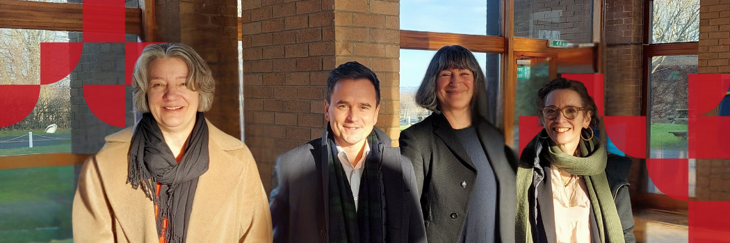 A group of three women and a man stand in front of some windows smiling at the camera