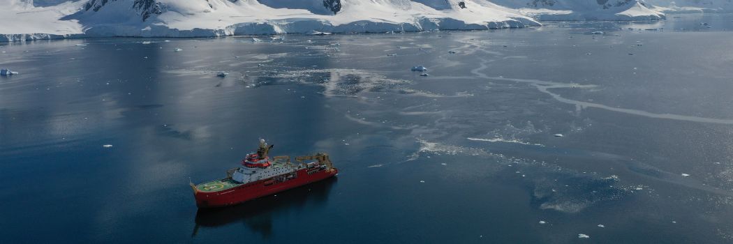 Aerial shot looking down on a boat on a blue, icy ocean with an ice sheet and snow capped mountains behind