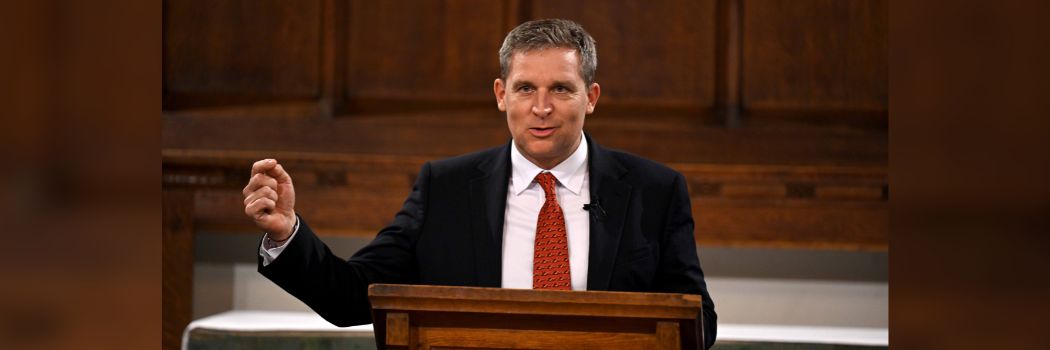 Male in black suite, white shirt and red tie standing behind lectern, speaking, right arm raised and pointing