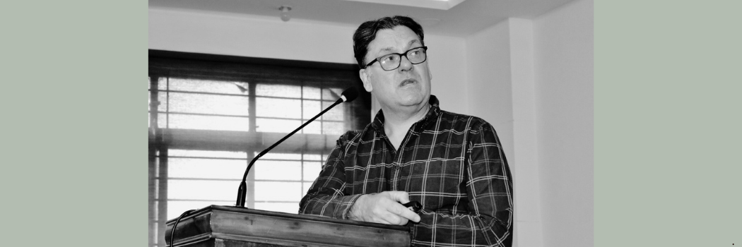 Black and white image of Professor Paul Denny at a lectern, looking over his shoulder towards the top right of the image