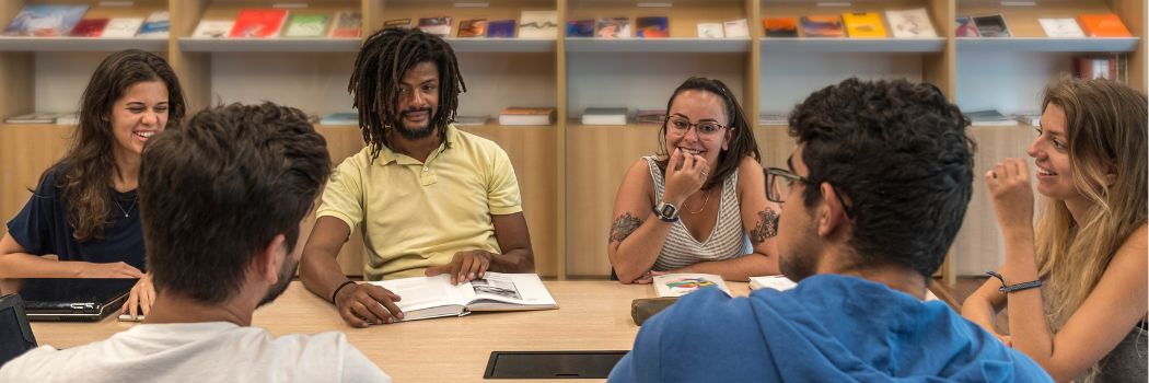 Six young adults sitting round a desk chatting, smiling and laughing. In the background are shelves of books