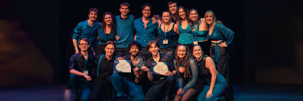 A mixed group of students wearing teal and black in a group holding two certificates and a trophy