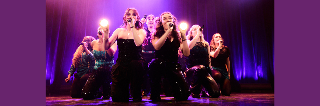 Members of Northern Lights A Cappella performing on stage with choreography suggesting a motobike and rider. The front two singers are holding headlamps.