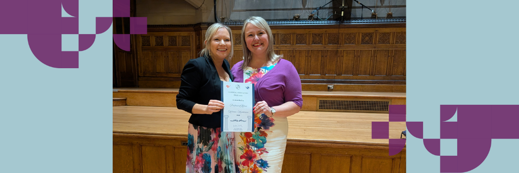 Two women wearing bright floral dresses smiling at the camera while holding a certificate.