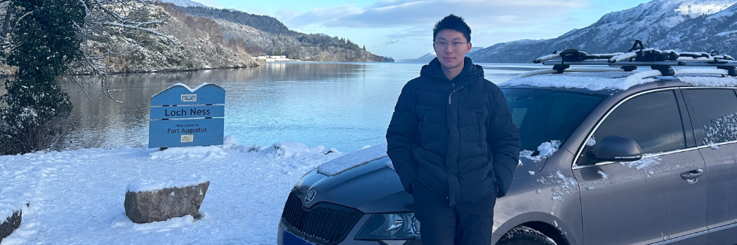 A student wearing warm clothing perching on a car bonnet near a sign for Loch Ness. The loch is visible in the background.