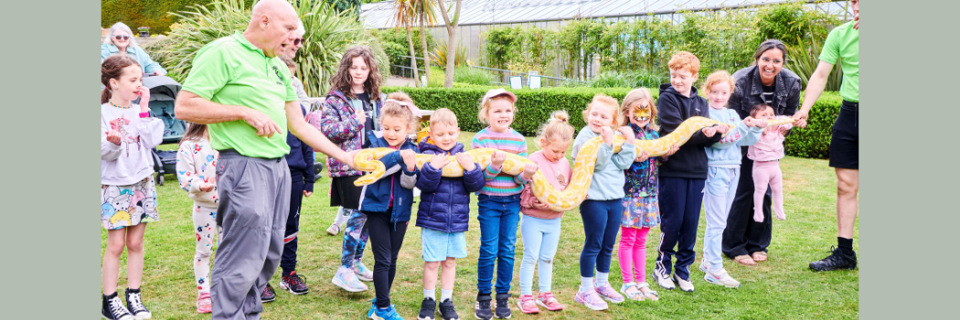 A group of children standing in a garden holding a large yellow snake between them.