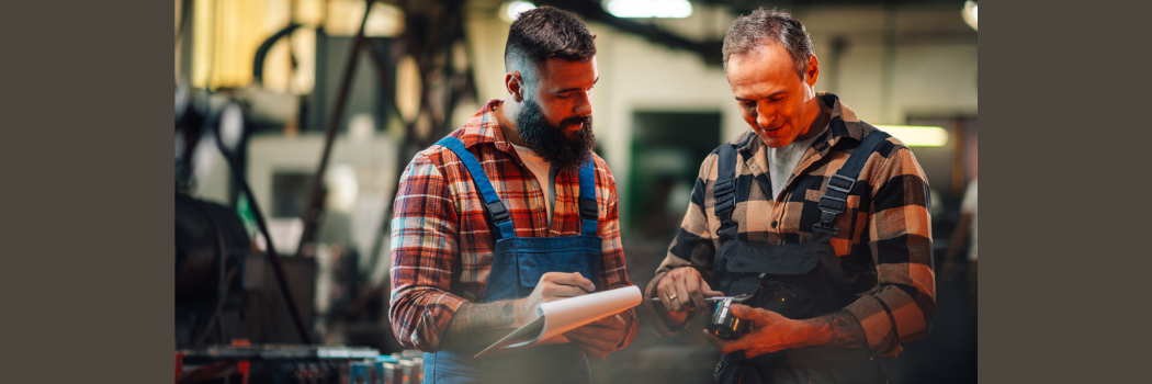 Two men in checked work shirts and blue overalls examining a metal object in a workshop.