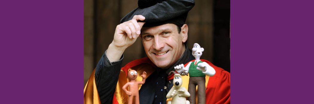 A man in wearing red academic robes and a black academic cap smiles at the camera. In front of him are models of the characters Morph and Wallace & Gromit