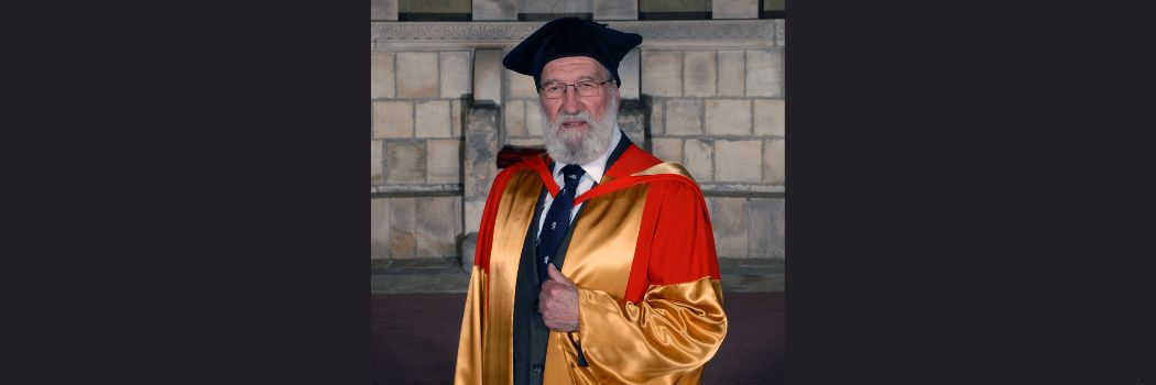 Dr Fenwick Lawson wears gold and red academic robes and a black academic cap while looking at the camera.