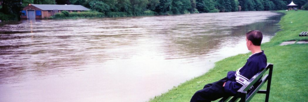 A man sits on a bench next to the flooded River Wear