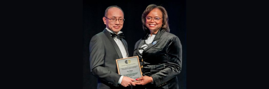 A man in a tuxedo and bow tie receives an award plaque from a woman in black velvet jacket.
