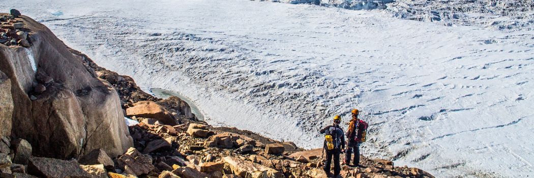 Two scientists standing on rocks overlooking the edge of the icy, white, Mawson Glacier, East Antarctica.
