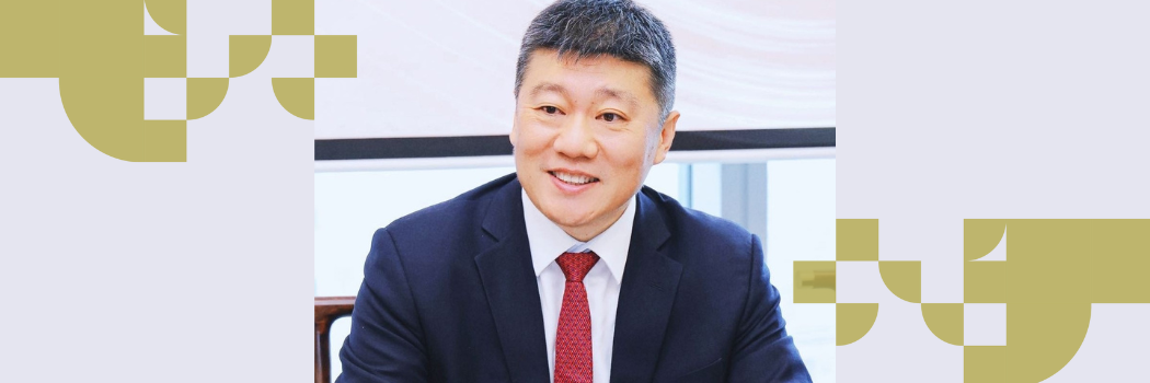 A man with short greying hair smiling while wearing a suit, white shirt and red tie.