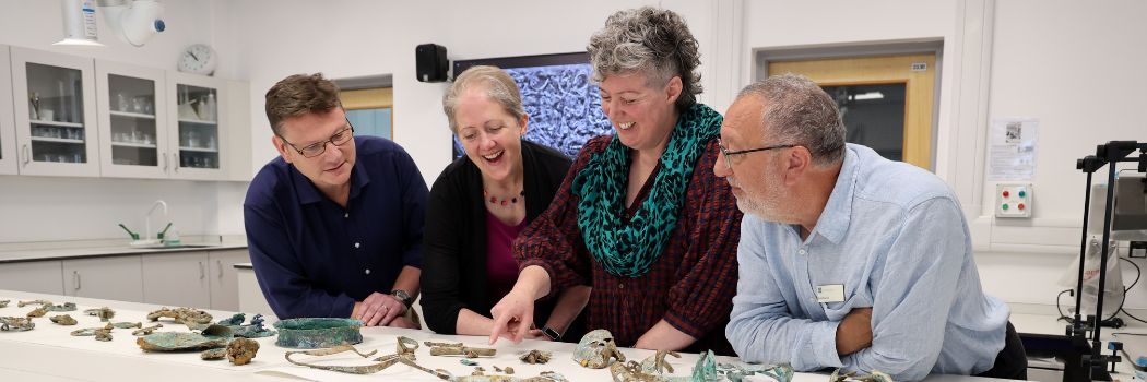 A group of four people stand behind a white counter top in a lab with a range or archaeological objects in front of them