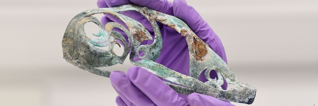 An intricate piece of metal work is held in a person's hands. The person is wearing purple latex gloves.