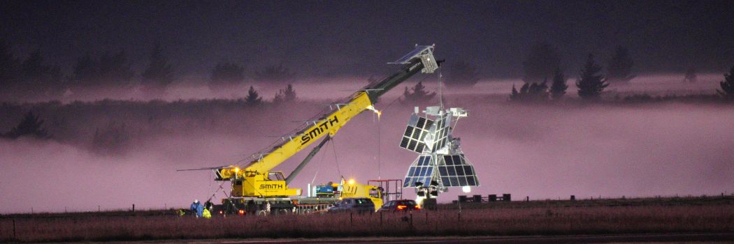 The SuperBIT space telescope being hoisted by a yellow crane against a black and pink dusk sky