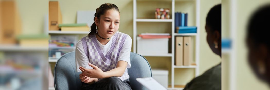 A young woman sits in a chair facing the camera, while talking to a therapist.