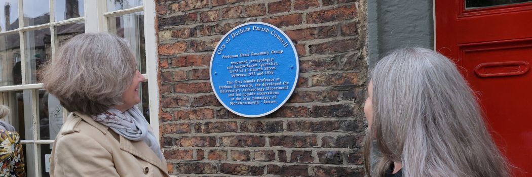 Professors Karen O'Brien and Sarah Semple look at a round blue plaque, set against a red brick wall, in honour of Professor Dame Rosemary Cramp.