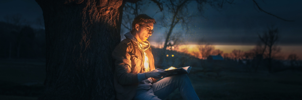 A man in a jacket sits under a tree in the dark reading a book, with a light illuminating his face