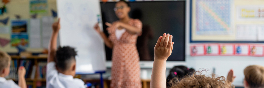 A child with their hand raised in a classroom with a teacher in the background in front of a blackboard