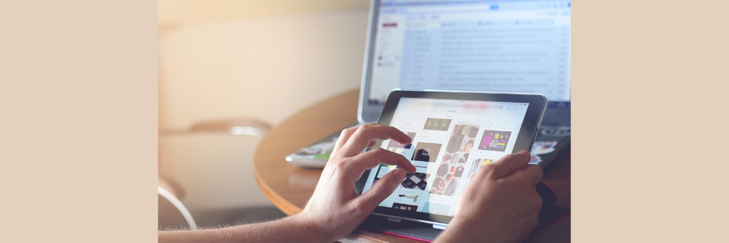A close up of a woman's hand scrolling on a tablet with a computer screen in the background