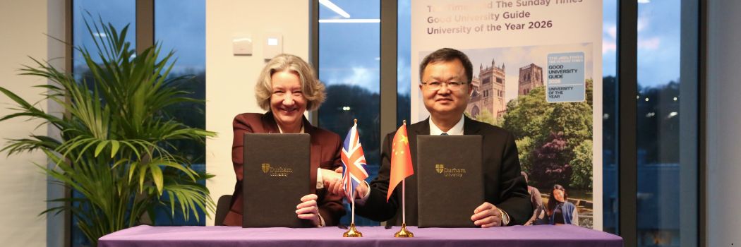 Two people sit behind a desk shaking hands while holding leatherbound document folders. On the desk are the flags of the UK and China.