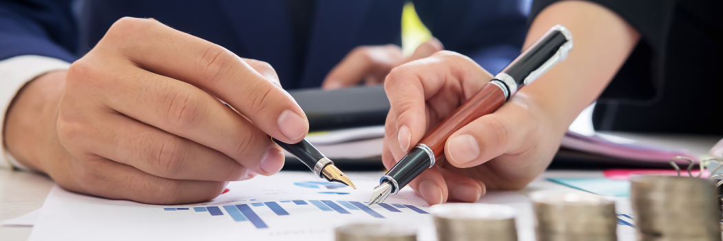 Two hands holding fountain pens above a paper containing graphs with coins sitting on the table