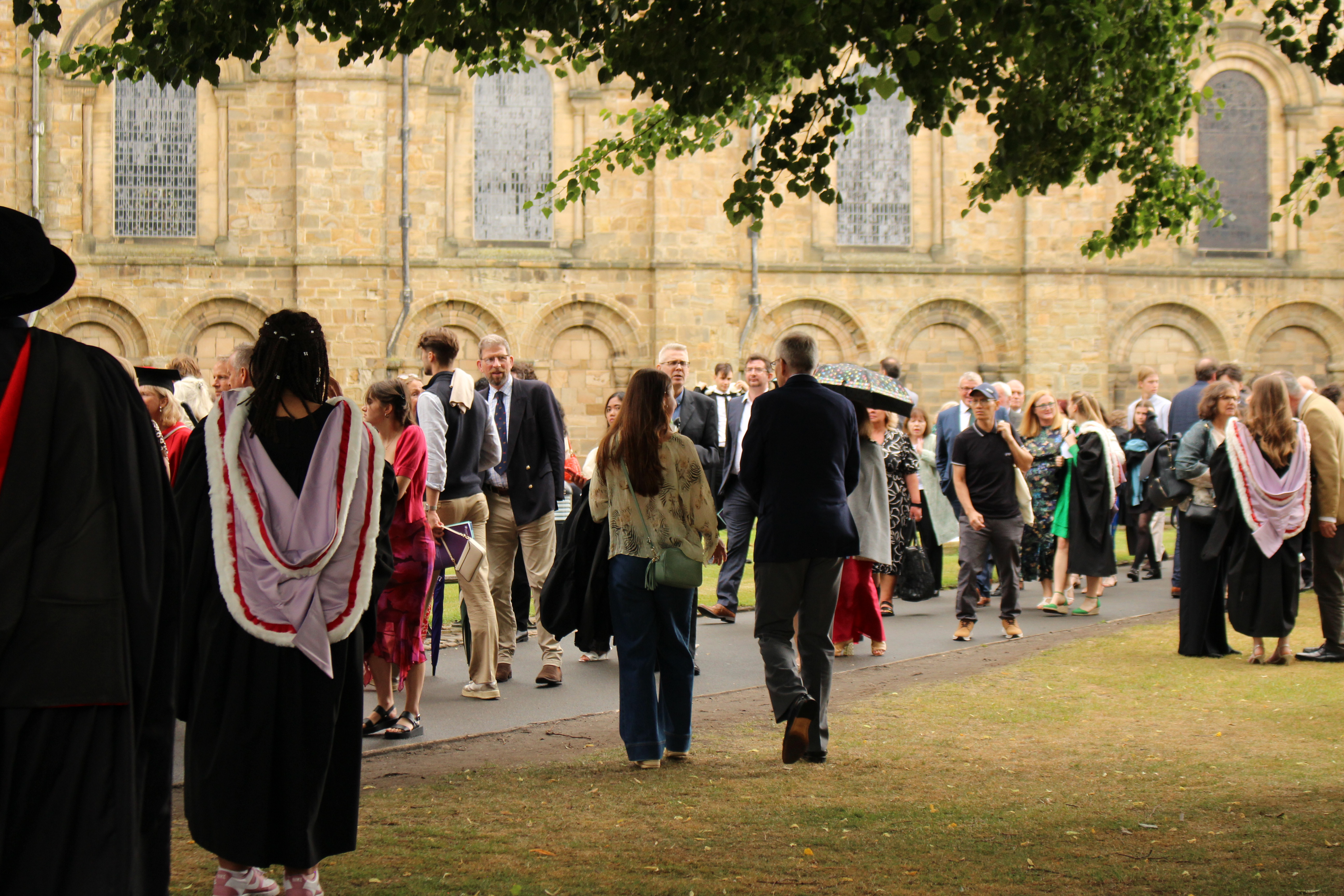 Graduates and their families gather outside Durham Cathedral for a graduation ceremony. People dressed in graduation robes and colourful hoods mingle with guests in formal attire along a tree-lined path. The backdrop is the historic Cathedral, with its arched windows and Romanesque architecture, creating a celebratory and picturesque atmosphere.