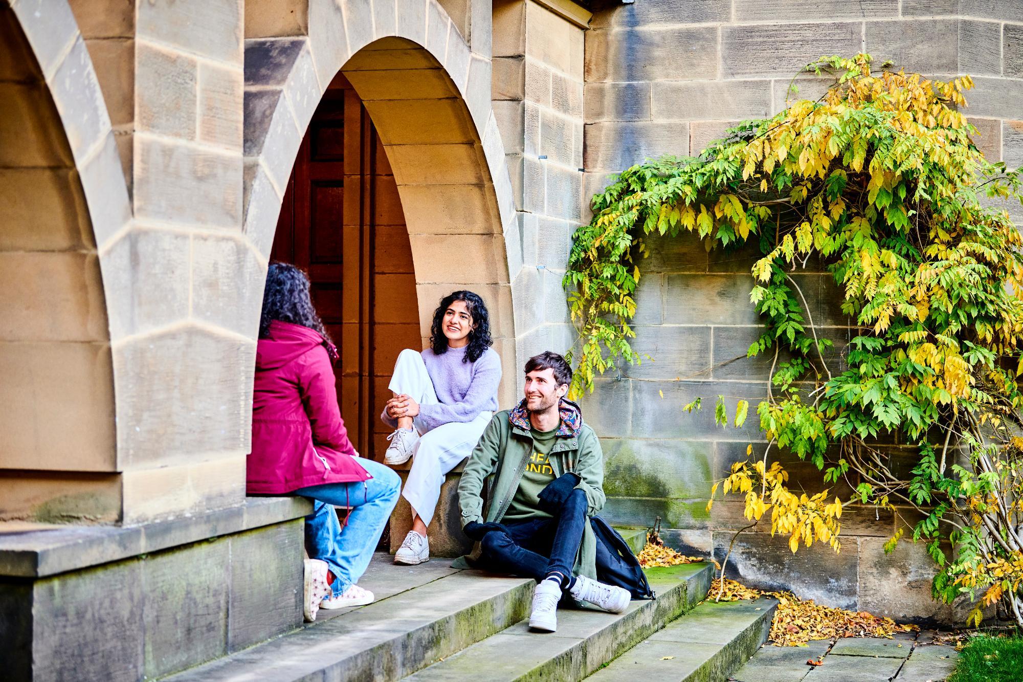 Three students sitting in the courtyard against limestone buildings on a sunny day talking socially