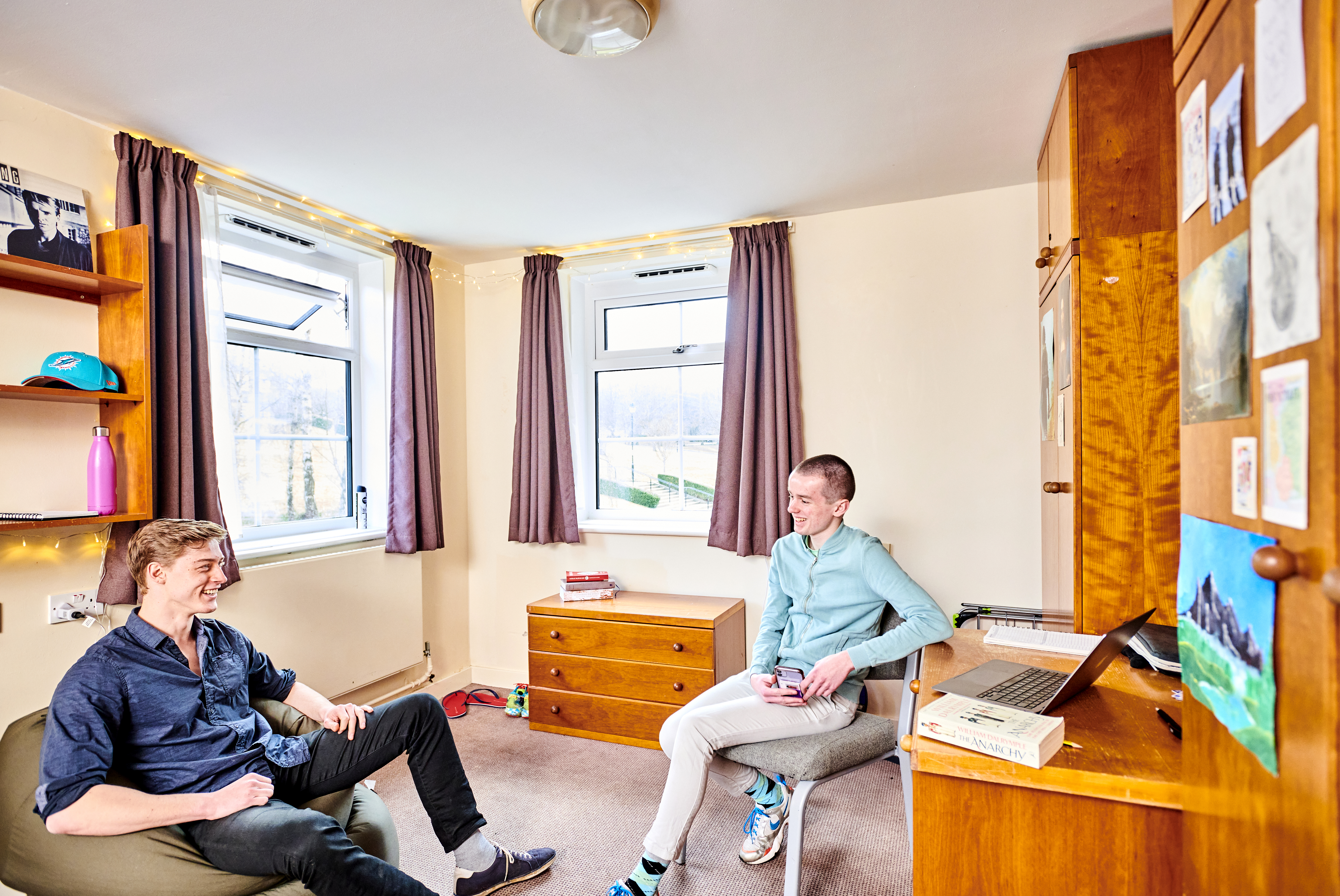 two students sitting in a bedroom, one in a chair and one on a beanbag, having a friendly conversation while relaxing