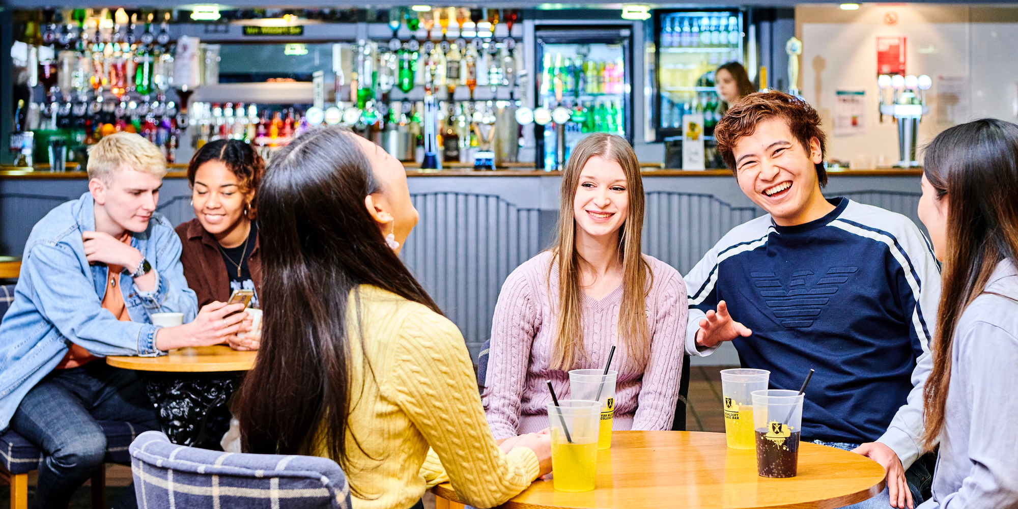 A group of students having a drink while seated around a table while several other students sit and talk at another table