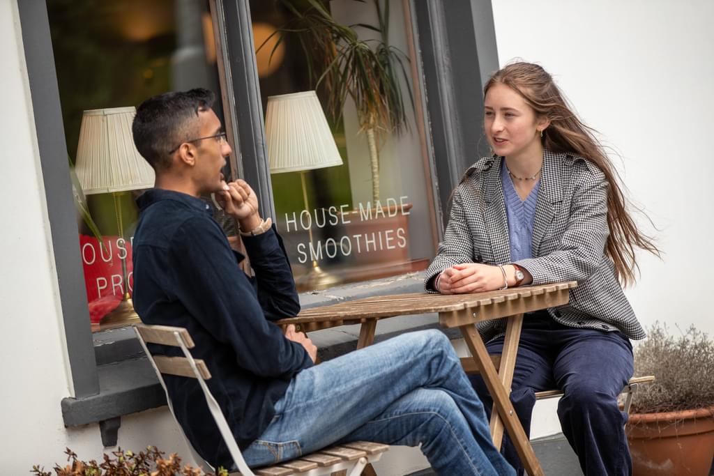 A young man and woman sit opposite each other at a round wooden table. They are sitting outside Whitechurch cafe.