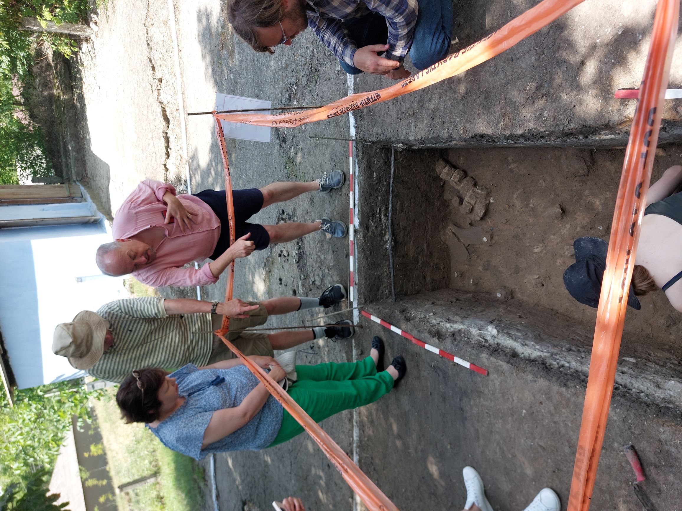 Three people stand by an archaeological trench bordered by orange cordons. They are Professor Paul Pettitt, Ambassador Fern Horine, and her husband David. The trench contains 25000 year old campsite elements including an articulated mammoth spinal column. A dark blue hat is also visible at the bottom of the photo.