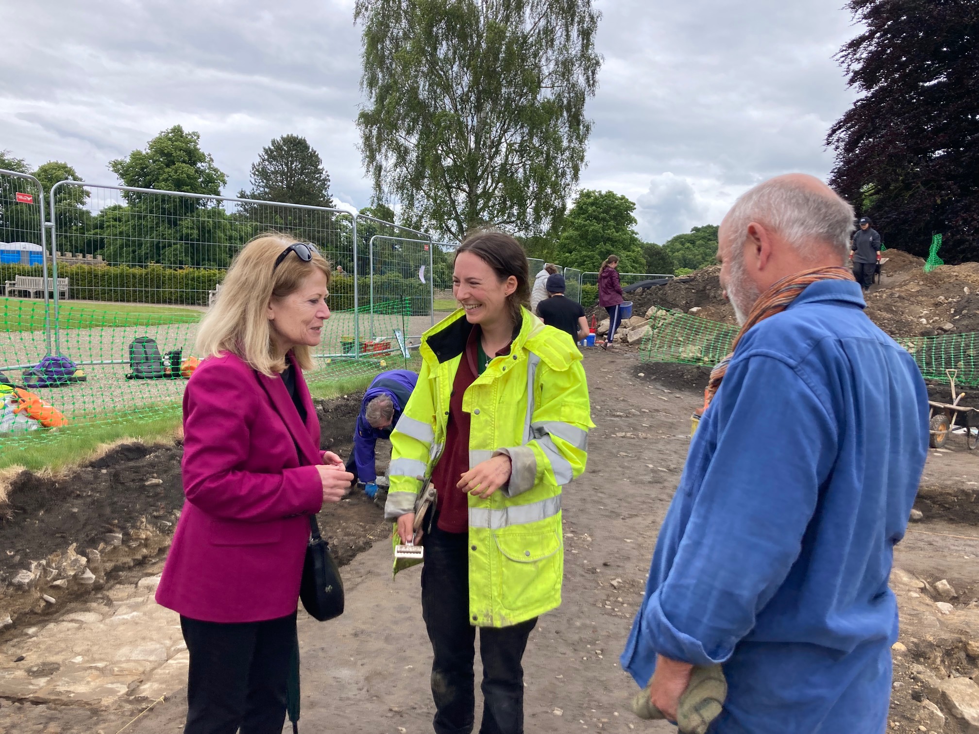 Three people stand in an archaeological trench having positive conversation. The student in the middle is smiling broadly and wears a high-vis jacket. Other students are in the background with buckets and trowels excavating within the trench.