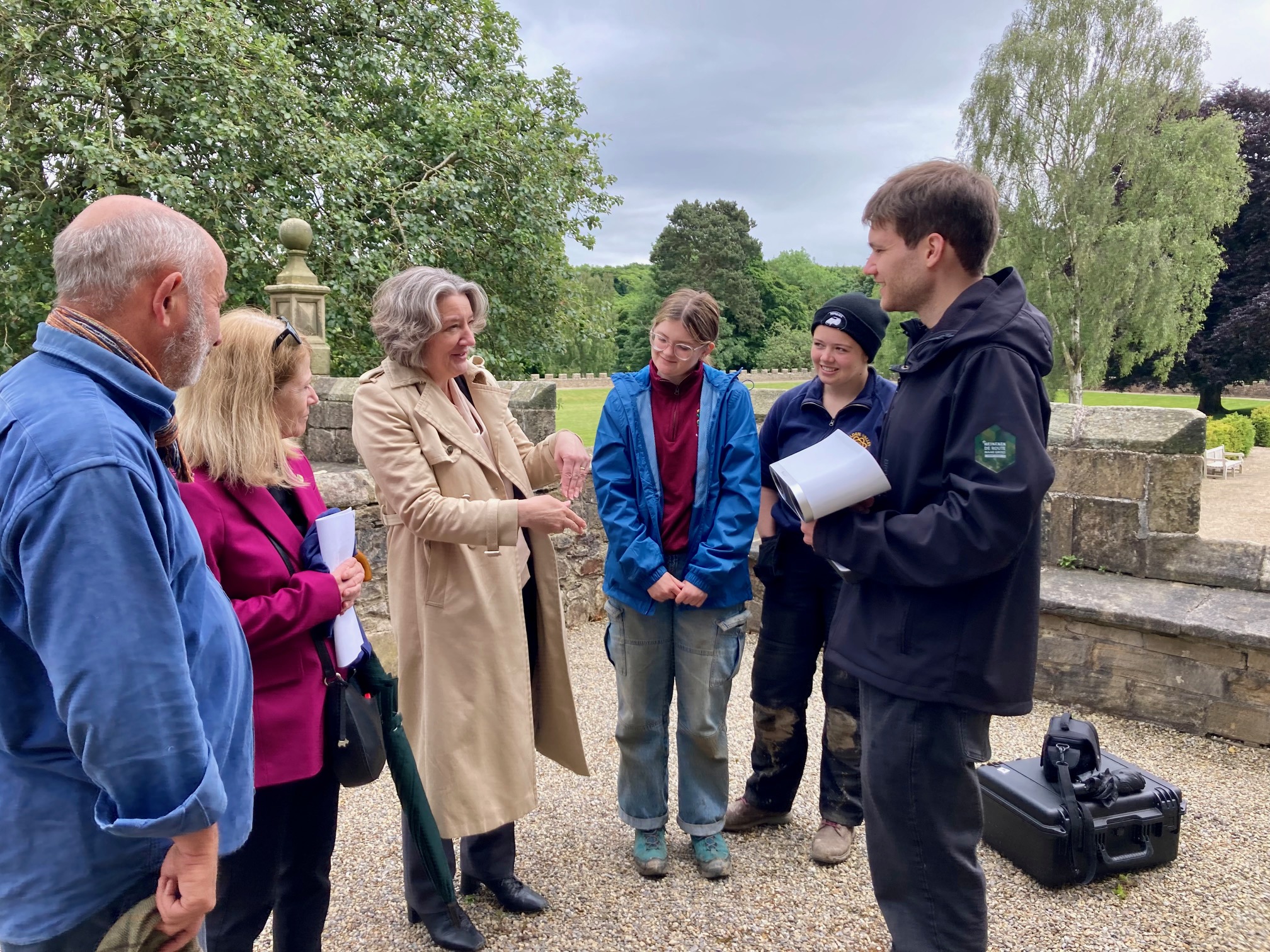 Six people standing together having a discussion near some low medieval stone walls. Two are Archaeology staff members, two are Archaeology students and two are members of senior Durham University leadership.