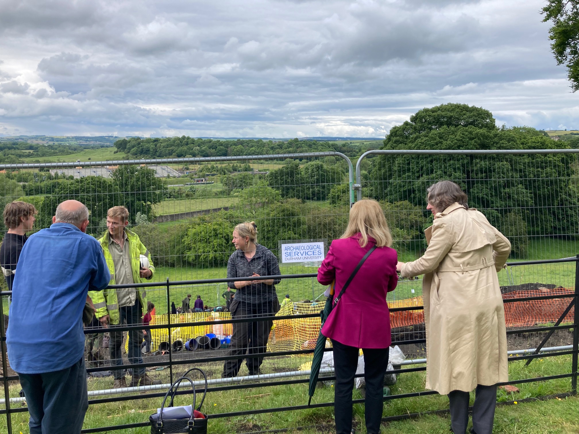 Three people stand the near side of a metal fence on a grassy slope, facing away from the camera. They are speaking to three people on the other side of the fence, one of whom is wearing a high-vis jacket and holding a helmet. A sign on the fence reads Archaeological Services Durham University. Downhill, there are some yellow plastic mesh fencing and buckets visible.