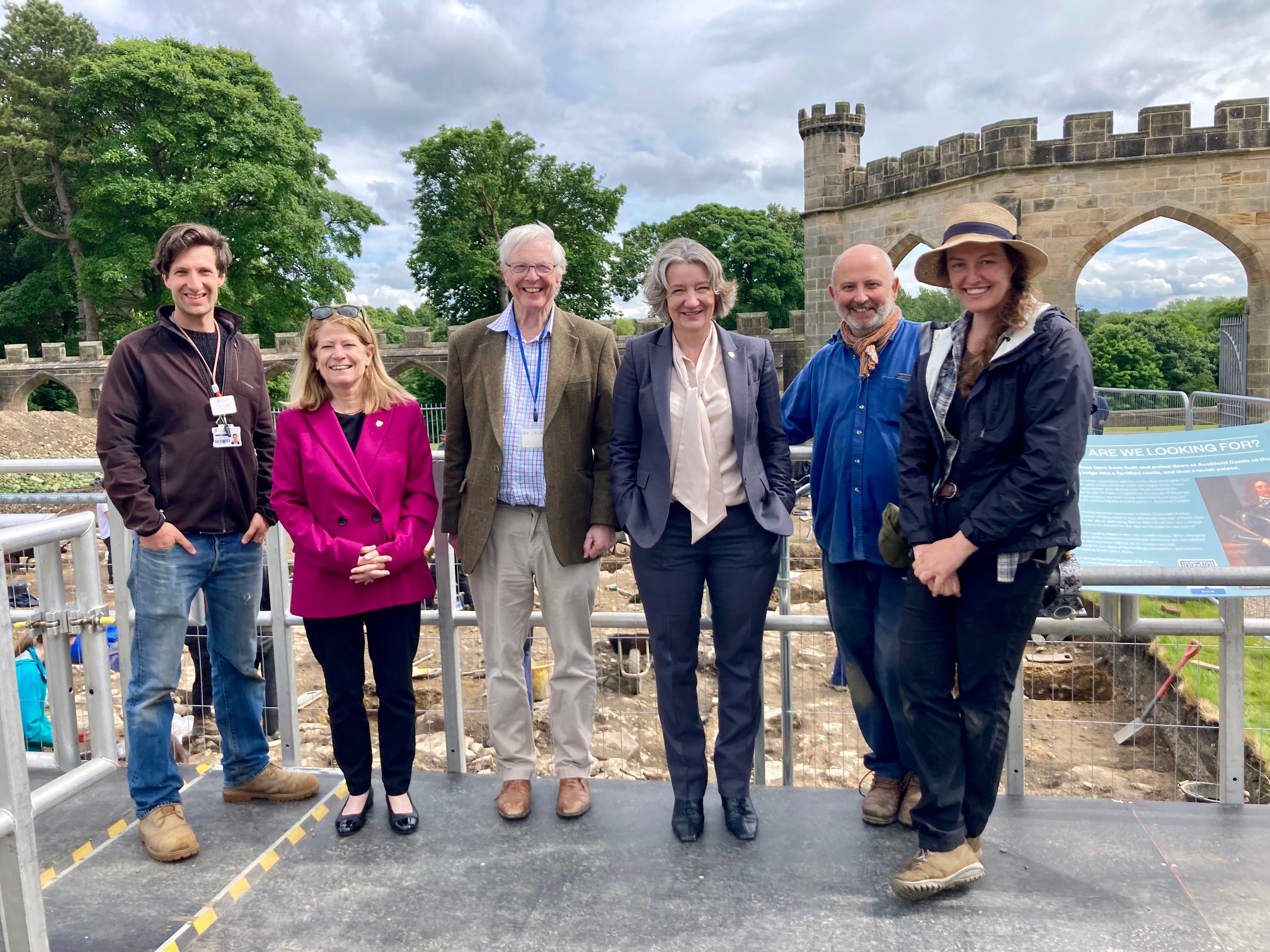 Six people smiling at the camera, stood on a sunny viewing platform overlooking an archaeological trench. In the background are some medieval arched stone walls and trees, under a bluish grey cloudy sky.