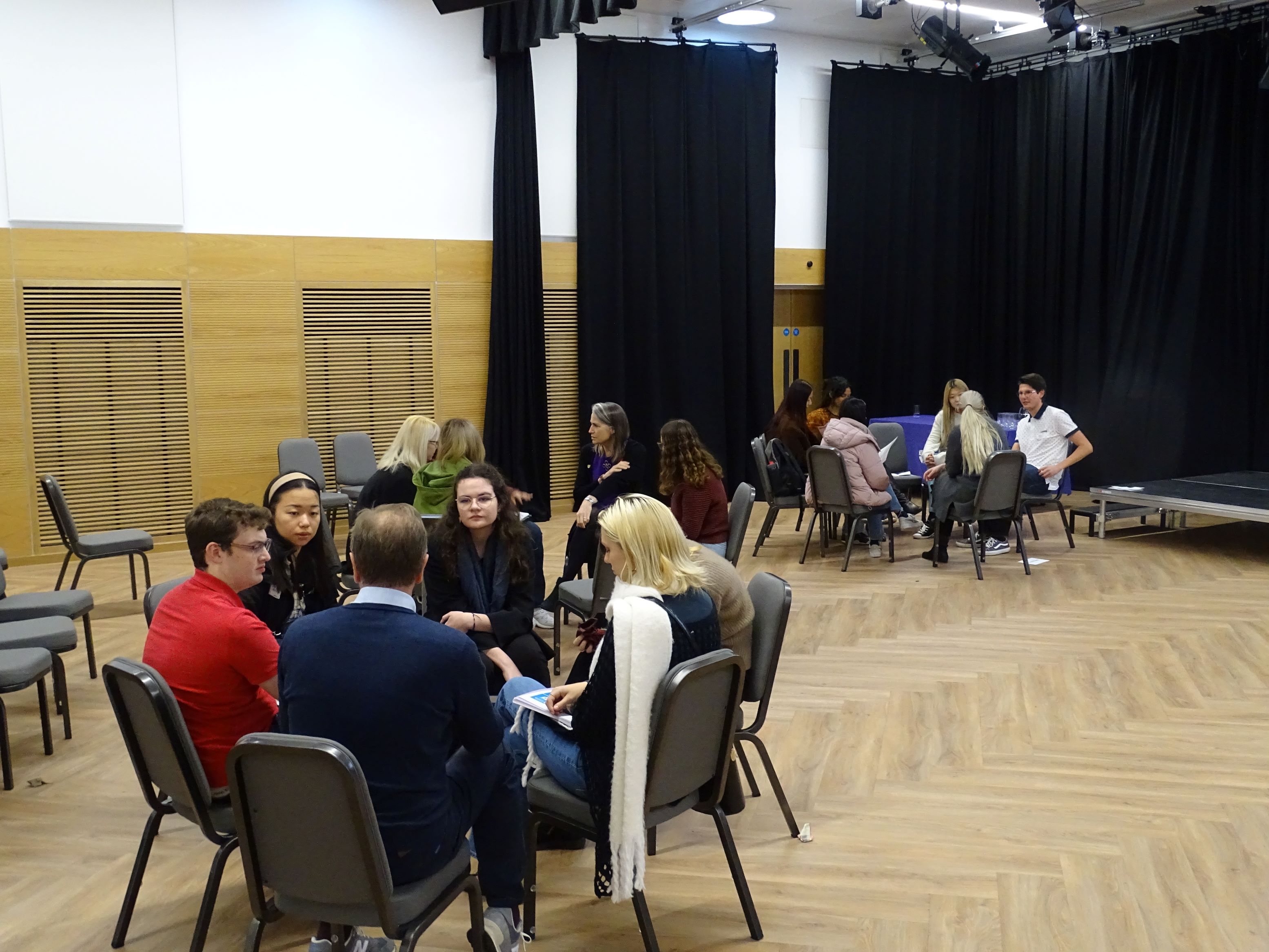 Groups of people sat in discussion circles in a wooden floored auditorium with black stage curtains.