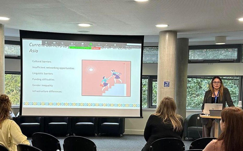 Durham PhD Student Aiya Raissova standing at a table next to a screen, giving a talk on the current barriers that women in archaeology in Central Asia face.