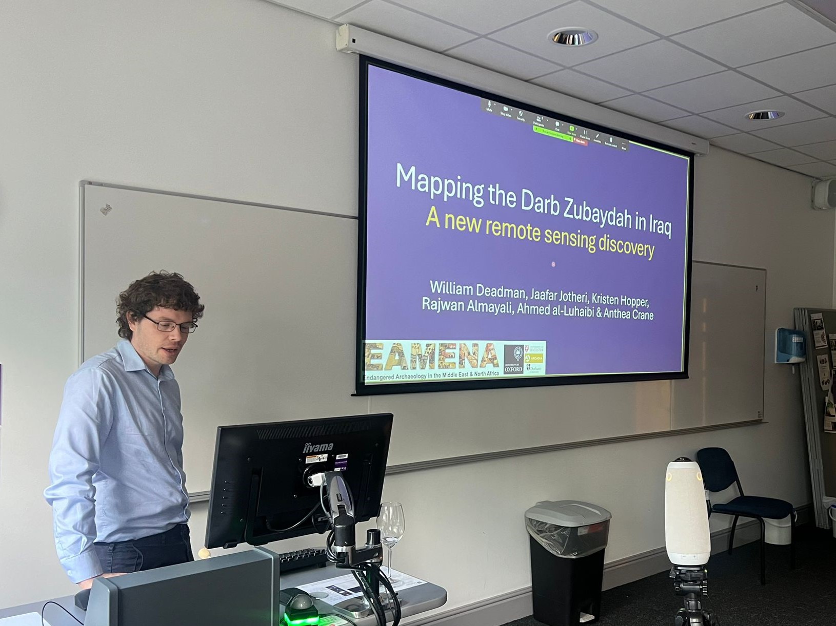 William Deadman standing behind the lecture desk, with the presentation titled 'Mapping the Darb Zubaydah in Iraq: A new remote sensing discovery.