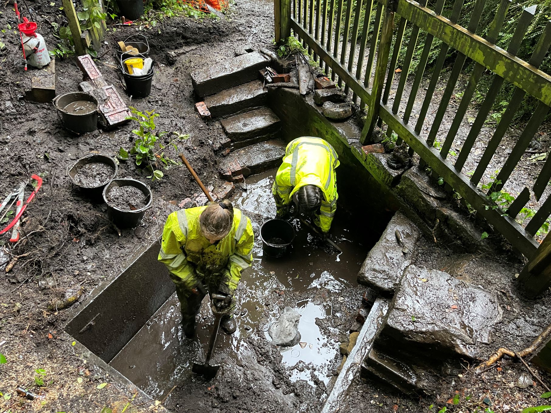 Two archaeologists in high-vis jackets and waterproof attire standing in a wet and muddy trench next to some steps and a fence.