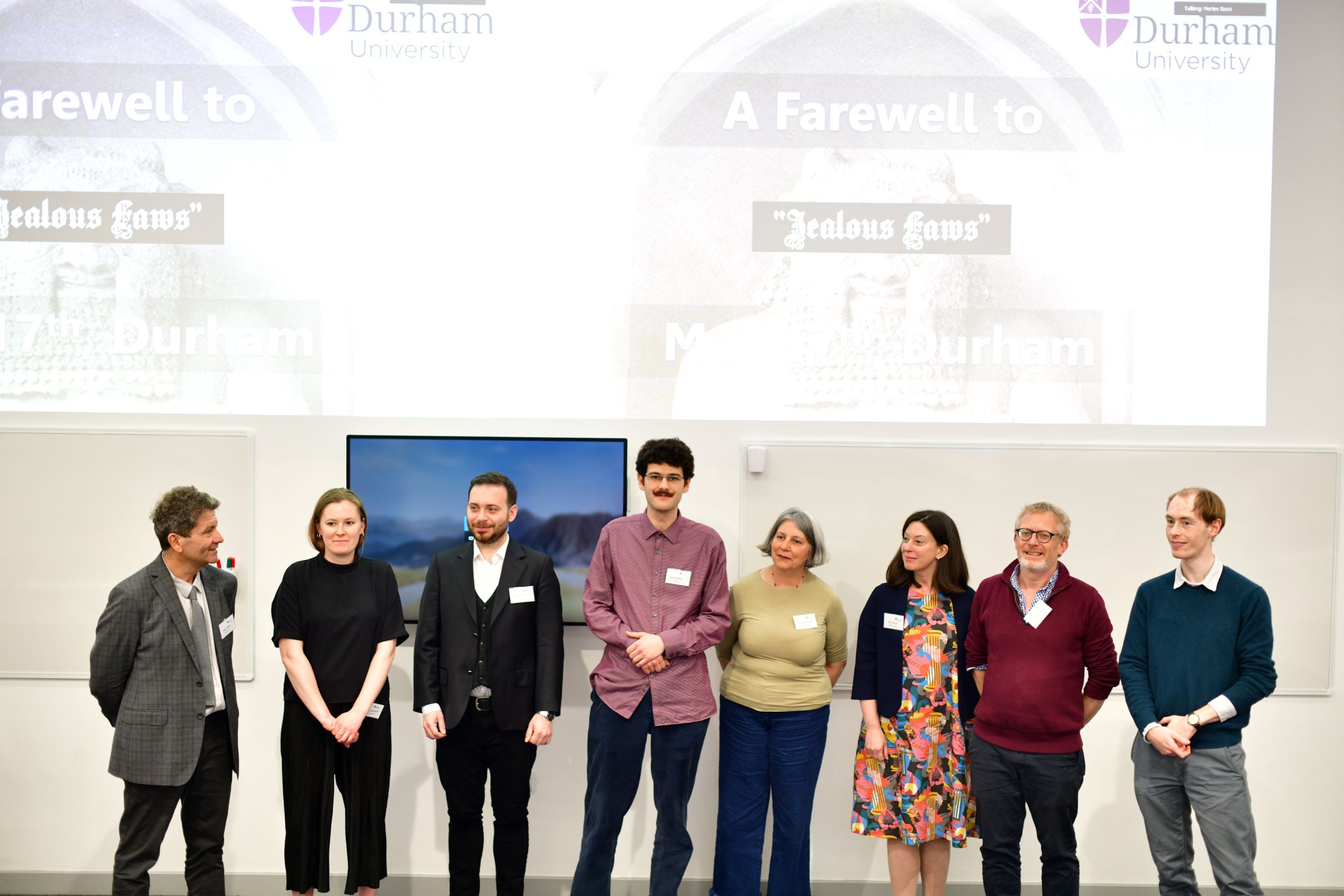 Eight people stand in front of two screens and whiteboards, at the front of a lecture theatre.