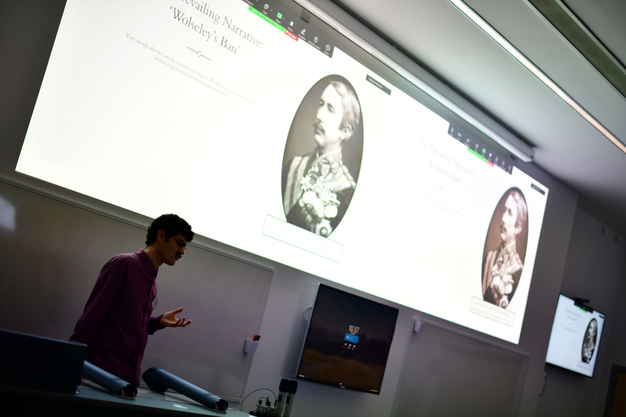 Pertev Basri stands at the front of a darkened lecture theatre presenting to an out-of-frame lecture theatre audience.