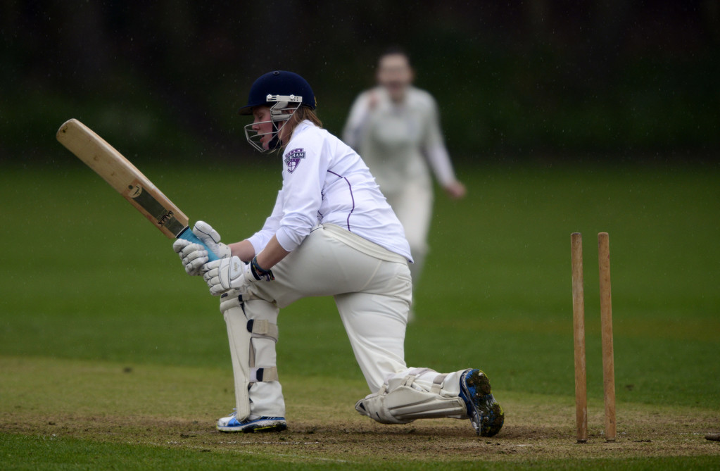 Female cricketer holding bat on one knee in front of wickets. One stump is missing