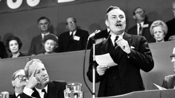 A man stands at a microphone with a piece of paper in his hand delivering a speech. People can be seen sitting behind him listening.